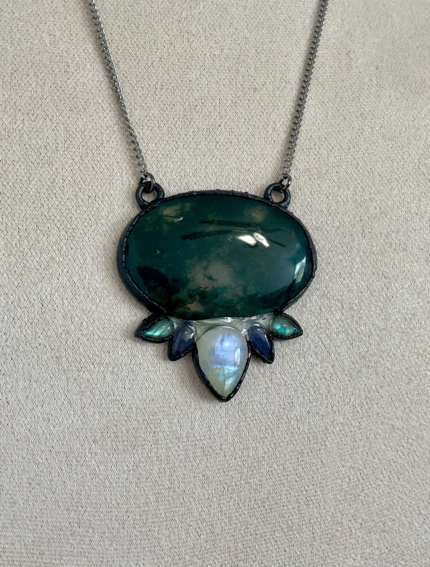 Close-up of a green moss agate pendant accented with rainbow moonstone and labradorite, forming a lotus-like design in a dark gunmetal setting.