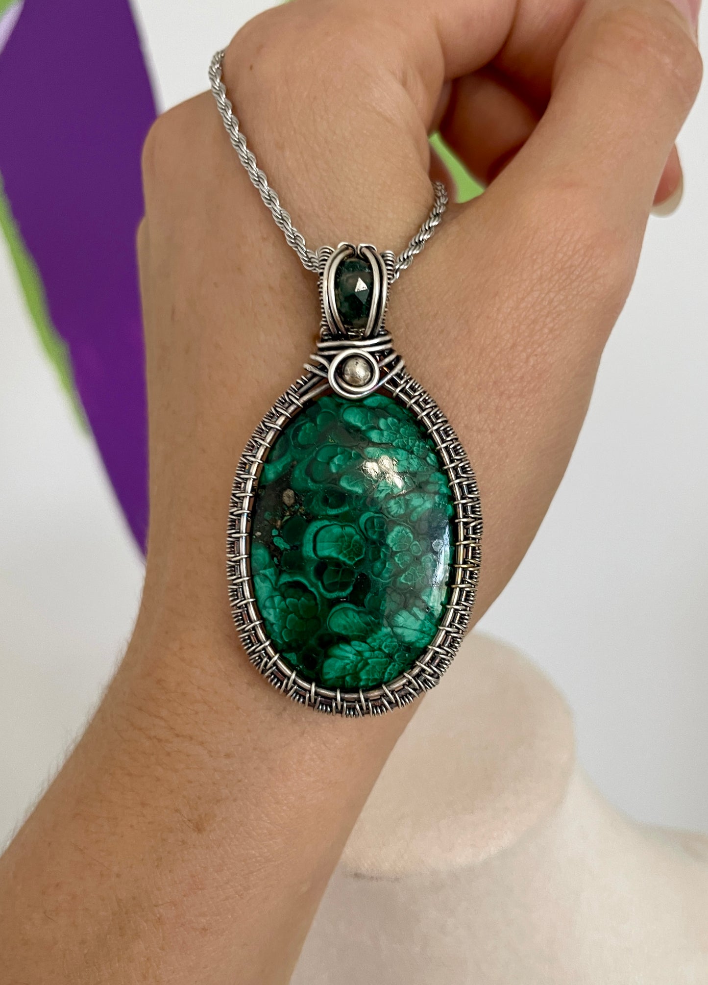 Hand holding a large oval malachite gemstone pendant with a rare plume pattern, wrapped in detailed .925 sterling silver wirework, featuring a green moss agate accent and stainless steel chain.