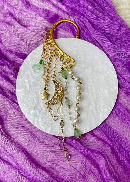 Gold ear cuff adorned with textured crescent moon charm, green glass flower, starry chain, green aventurine chain, glass pearls, and star charm, inspired by natural elegance and celestial allure.