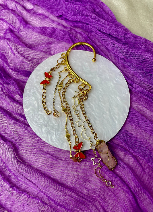 Gold ear cuff adorned with textured cactus charms, sun and moon chain, red evil eye butterfly charms, iridescent glass beads, and hand-wrapped iron-stained quartz piece, inspired by desert landscapes and celestial beauty.