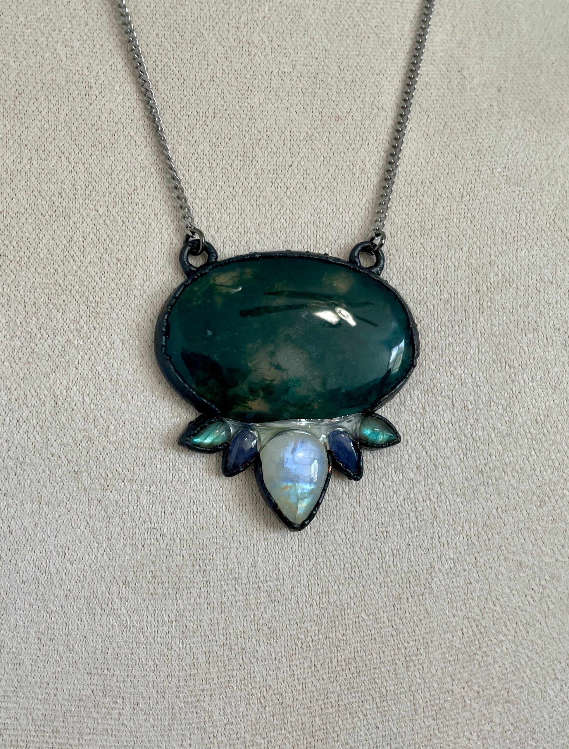 Close-up of a green moss agate pendant accented with rainbow moonstone and labradorite, forming a lotus-like design in a dark gunmetal setting.